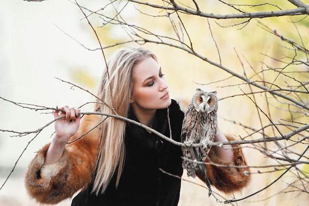 Красивая женщина в шубе с совой на руке. Блондинка с длинными волосами в природе держит сову. Романтический нежный образ девушки