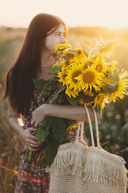 Beautiful woman gathering sunflowers in warm sunset light in summer meadow Tranquil atmospheric moment in countryside Stylish young female in floral dress holding sunflowers in evening field