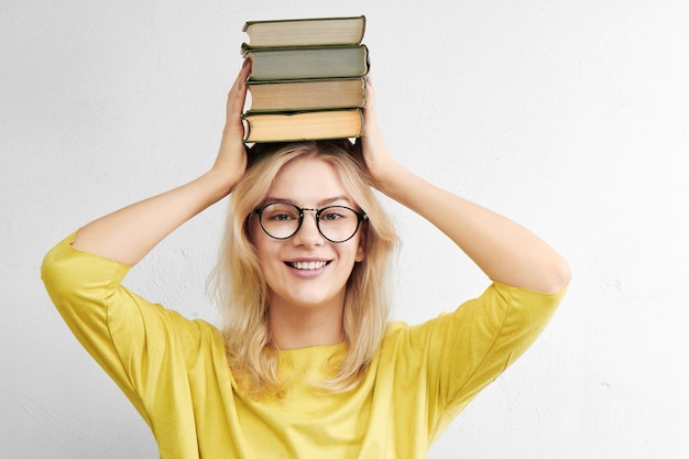 A beautiful woman in glasses is holding a stack of books above her head and is smiling toothyly on a white background. Happy and fun education