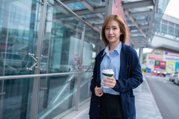 写真 街の通りで働く途中でホットドリンクのカップを手に持っている成功したビジネスウーマンの肖像画オフィスビルの近くを歩いてコーヒーと一緒に仕事に行く美しい女性高解像度