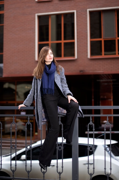 Красивая женщина в сером пальто и синем шарфе над зданием с окнами. Молодая женщина сидит на перилах и кокетливо смотрит