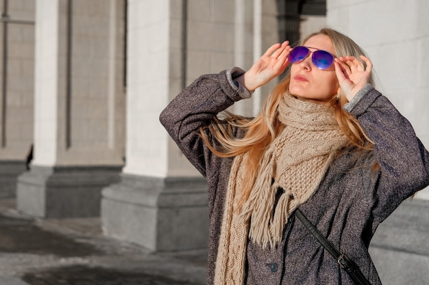 A beautiful woman in a gray coat and sunglasses walks on the street in the city.