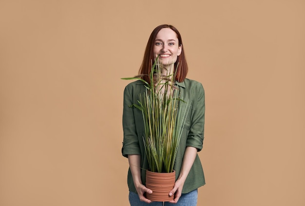 Beautiful woman in green casual shirt smiling broadly looking at camera carrying a potted houseplant
