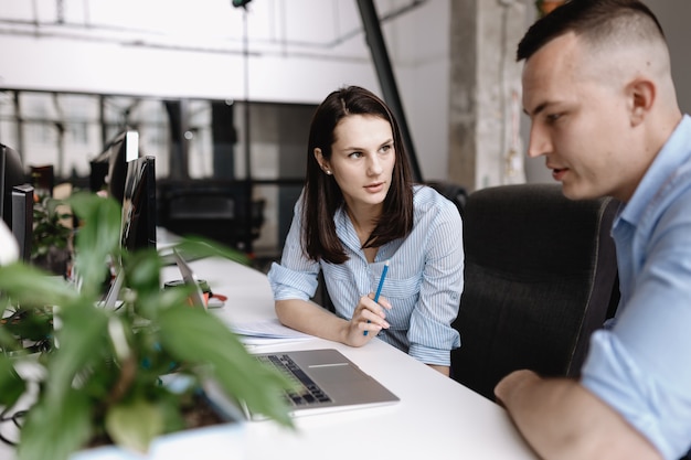 Beautiful woman and guy dressed in office style clothes are working together with laptop sitting at desks in the light modern office