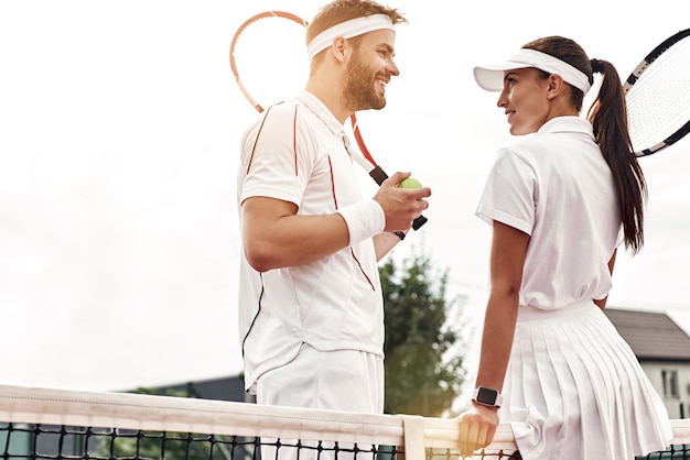 Beautiful woman and handsome man are going to play tennis.\
woman in white polo and skirt looks ar her partner. man in white\
sportswear with the racket and the ball in his hands looks at\
her