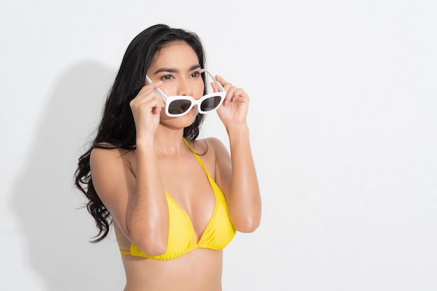 Bella donna nella felicità che indossa un abito bikini giallo e che tiene gli occhiali da sole bianchi post nel tiro in studio di moda estiva di concetto isolato su priorità bassa bianca.