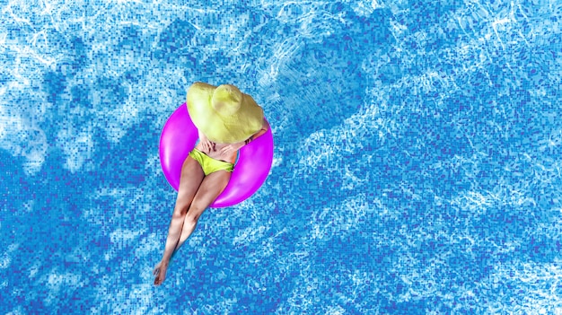 Beautiful woman in hat in swimming pool aerial top view from above, young girl in bikini relaxes and swims on inflatable ring donut and has fun in water on family vacation, tropical holiday resort