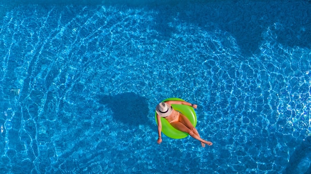 Beautiful woman in hat in swimming pool aerial view from above, young girl in bikini relaxes and swims on inflatable ring donut and has fun in water on vacation