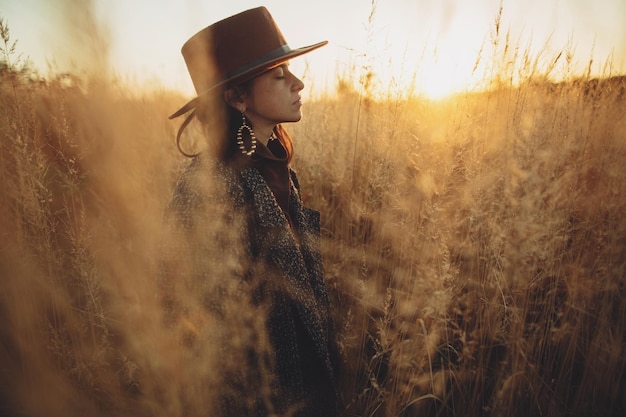 Foto bella donna in cappello e cappotto vintage in piedi in erba nel caldo tramonto in campo autunnale tranquillo