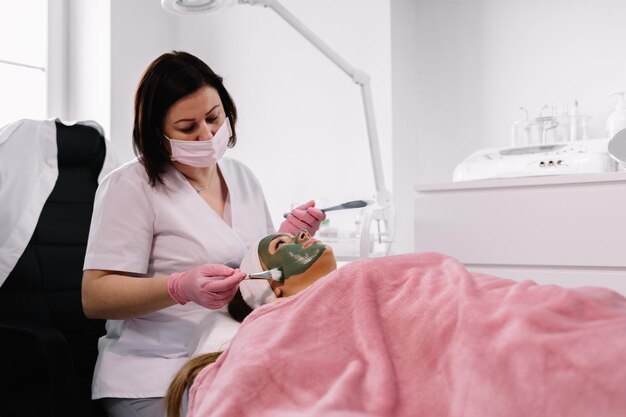 Beautiful woman having clay facial mask apply by beautician