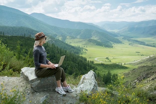 Beautiful woman hipster traveler freelancer with laptop  on the top of the mountain. Remote work concept.