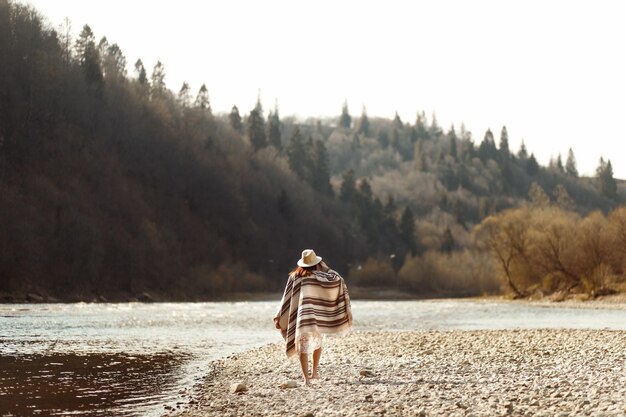 Beautiful woman hipster walking on river beach in mountainswearing hat and poncho boho travel concept space for text