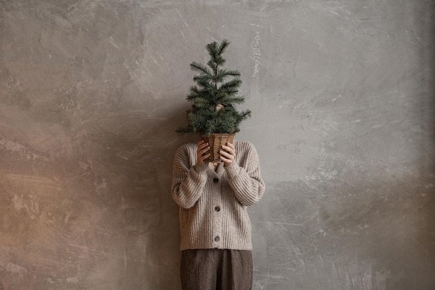 Beautiful woman holding bucket with fir needle branches against neutral concrete wall Aesthetic minimalist Christmas holiday composition