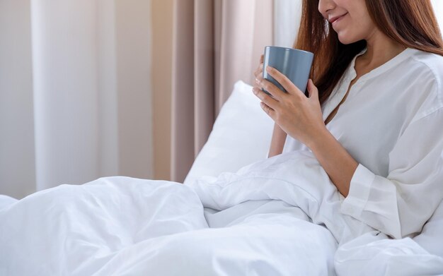 A beautiful woman holding and drinking coffee in a white cozy bed at home