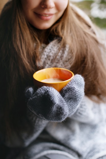 Bella donna che tiene e beve una tazza di caffè o cacao in guanti seduto a casa vicino alla finestra. sfondo sfocato albero di neve invernale.
