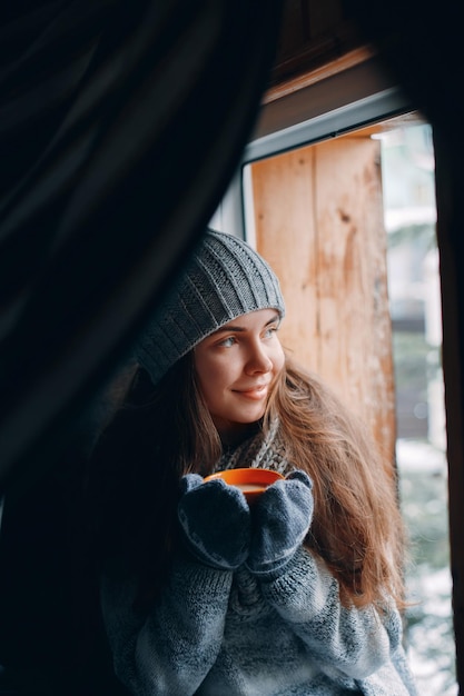 Beautiful woman holding and drinking a cup of coffee or cocoa in gloves sitting home by the window