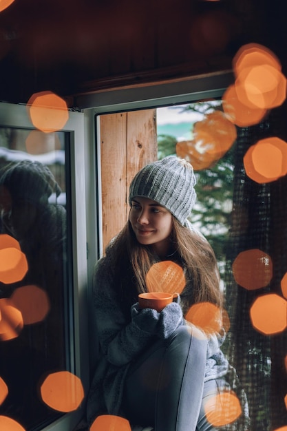 Beautiful woman holding and drinking a cup of coffee or cocoa in gloves sitting home by the window