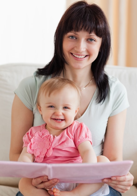 Beautiful woman holding her baby and a book in her arms while sitting on a sofa