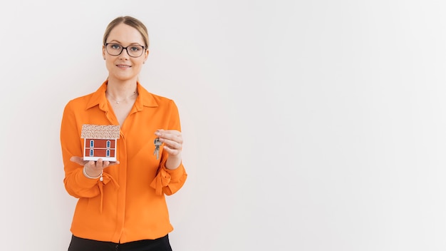 Beautiful woman holding house model and key in front of white wall