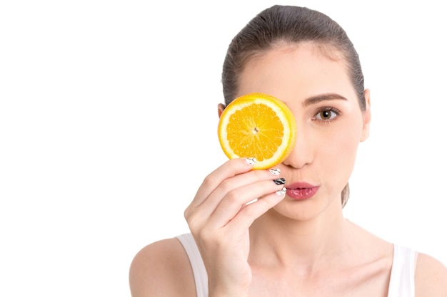 Beautiful woman holding orange slice hiding eye behind isolated on white background