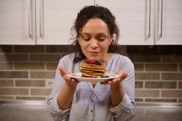 野生のベリーを添えておいしい自家製焼きたてのパンケーキのプレートを保持している美しい女性。 Shrovetideのために調理されたパンケーキを持つ魅力的な若い黒髪の巻き毛の主婦。パンケーキ火曜日