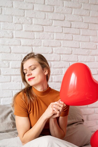 Beautiful woman holding red heart shape balloon sitting in bed