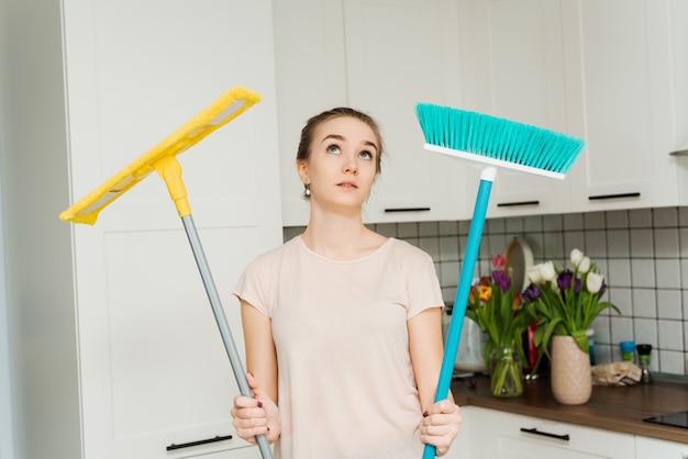 A beautiful woman holds a mop and brush for cleaning and mopping in her hands and sighs of fatigue. A housewife stands in the kitchen and wipes sweat from her face