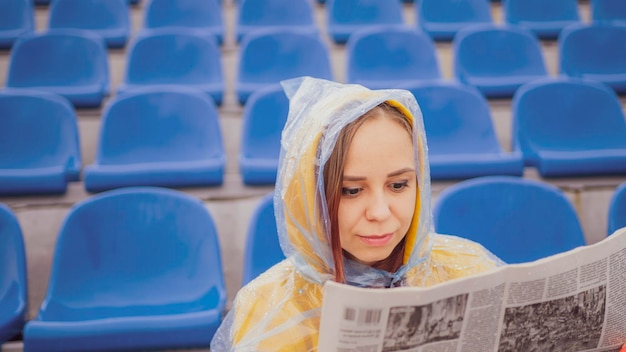 A beautiful woman in a hoodie and a plastic raincoat learns the latest news by reading a newspaper sitting on the podium in an empty stadium during the rain