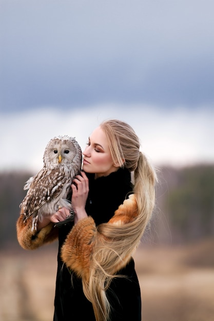 彼の腕にフクロウと毛皮のコートで美しい女性。フクロウを保持している自然の中で長い髪のブロンド