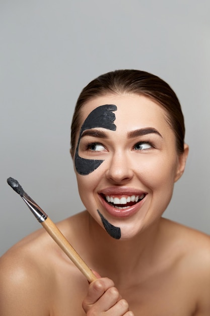 Beautiful woman is applying cosmetic spa black mask on face over grey background Beauty face