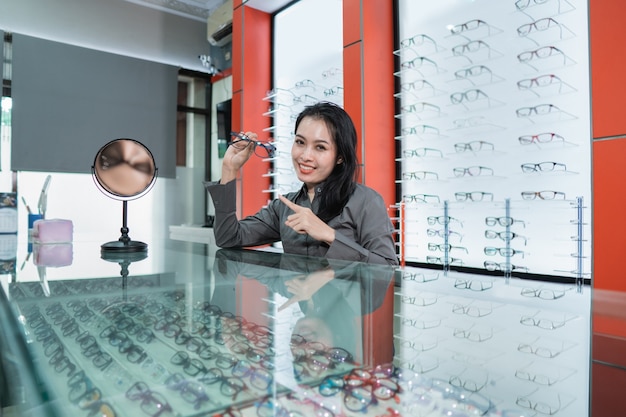 A beautiful woman is posing holding glasses against the background of a glasses display case in an eye clinic