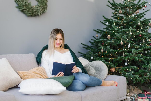 Beautiful woman is reading a book resting on pillows with a blanket at the Christmas tree at home