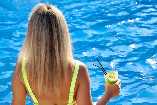 Beautiful woman is sitting next to the pool holding a glass with a drink