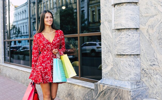 La bella donna sta camminando lungo la strada con un vestito floreale rosso, portando diverse borse della spesa, sorridendo e guardando avanti
