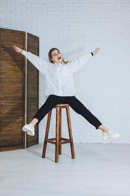 A beautiful woman is wearing a white cotton shirt and black pants Cheerful emotional girl sits on a wooden chair against a white brick wall