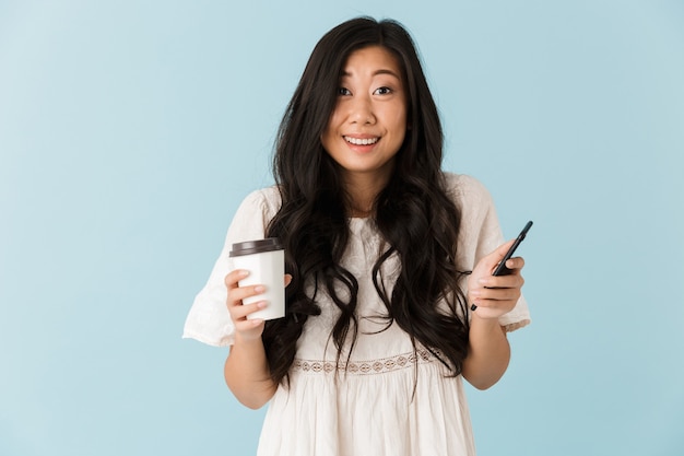 Beautiful woman isolated over blue wall holding coffee chatting by phone