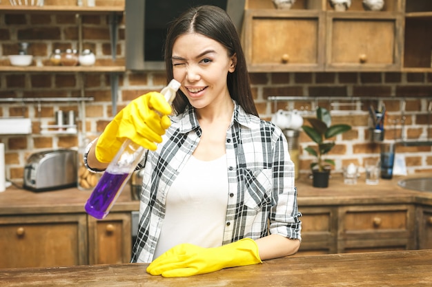 Красивая женщина на кухне улыбается и вытирая пыль с помощью спрея и тряпкой во время уборки ее дома, крупным планом