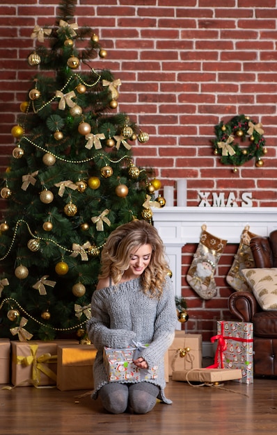 Beautiful woman in knitted dress sits on her lap and holds a New Year's gift in her hands.