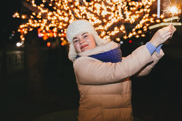 Beautiful woman in a knitted hat and scarf standing in the city with a sparkler. Celebration and Christmas concept.