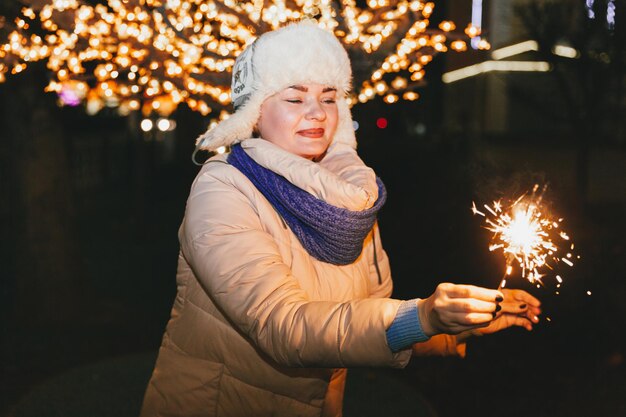 Beautiful woman in a knitted hat and scarf standing in the city with a sparkler. Celebration and Christmas concept.