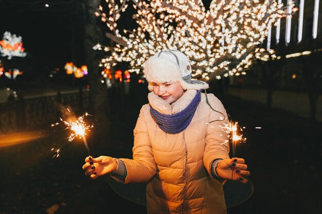 Beautiful woman in a knitted hat and scarf standing in the city with a sparkler. Celebration and Christmas concept.