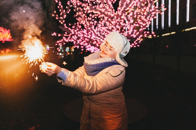 線香花火で街に立っているニット帽とスカーフの美しい女性。お祝いとクリスマスのコンセプト。