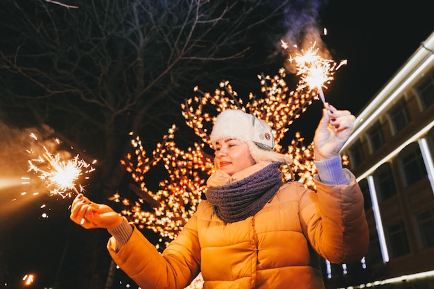 Bella donna in un berretto lavorato a maglia e sciarpa in piedi in città con una stella filante. celebrazione e concetto di natale.