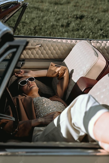 Photo a beautiful woman lies in a convertible in a field dressed in a beige jacket and white trousers and black sunglasses beautiful and stylish look