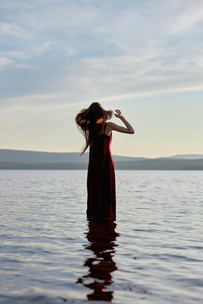 La bella donna in vestito rosso chiaro sta in acqua del mare del lago al tramonto. riflessione della ragazza e del cielo nell'acqua