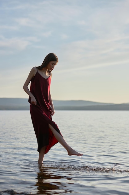 Beautiful woman in light red dress stands in water of lake sea at sunset. Reflection of the girl and sky in water