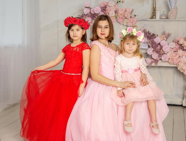 A beautiful woman in a long pink dress in a hall with flowers is photographed with her daughters who have wreaths of flowers on their heads The girls are cute Spring PHOTOSESSION Family