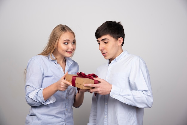 Beautiful woman looking at her boyfriend and giving him a present on gray wall.