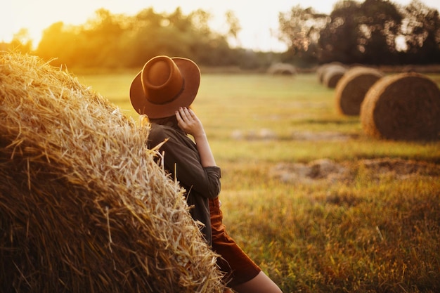 Photo beautiful woman looking at sunset at haystack atmospheric tranquil moment stylish girl relaxing at hay bale in summer field in evening sunshine countryside slow life