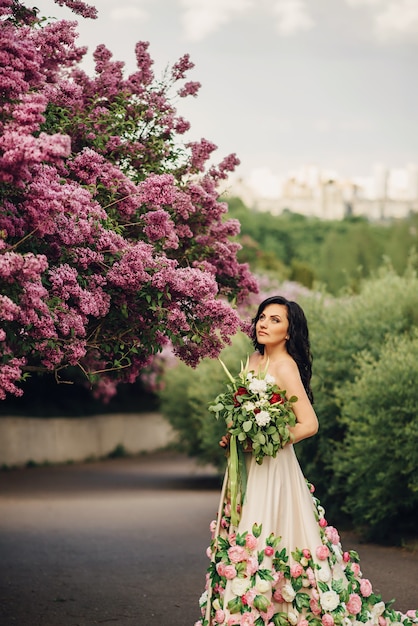 Beautiful woman in a luxurious dress is standing in a blooming lilac garden